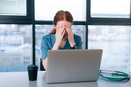 overworked young female doctor in blue medical uniform typing on laptop and rubbing eyes sitting at...