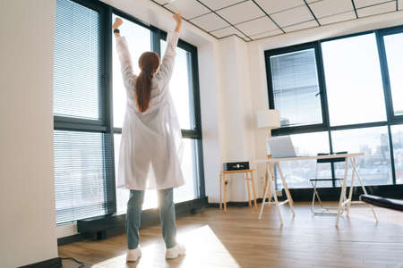 Back view of unrecognizable female doctor wearing white coat stretching hands on background of...