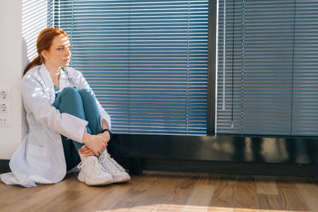Depressed young female doctor wearing white coat sitting on floor hugging knees near window with love in light medical clinic office room. Unhappy woman physician depression feeling sad at hospital.の写真素材