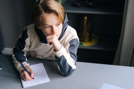 Close-up hands of unrecognizable pupil boy writing in paper notebook solving math equations sitting at desk in dark room. Back view of schoolboy doing homework at home.の写真素材