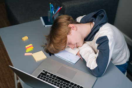Close-up of exhausted pupil boy sleeping on workbook in front of laptop after online lesson via Internet near window. Concept of distance education at home, closeup.の写真素材