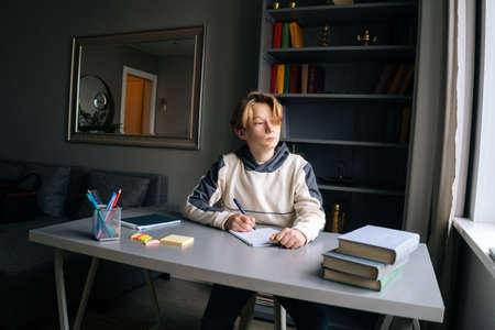 Portrait of thoughtful pupil boy looking away during making notes in copybook sitting at desk in children room. Child schoolboy doing homework at home. Concept of remote distance education.の写真素材