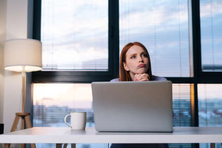 Front view of concerned redhead woman working on laptop computer looking away thinking solving...