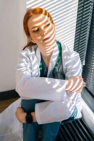 Close-up of desperated sad young female doctor in white coat sitting on floor hugging legs with hands near window. Unhappy woman physician depression feeling worried about professional malpractice.の写真素材