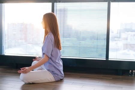 Young redhead woman meditating practicing breathing yoga exercise, sitting on floor near window in...