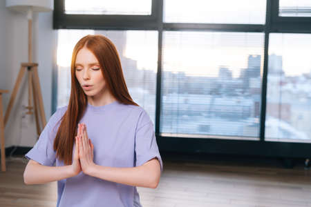 Close-up of redhead focused young woman meditating sitting on floor in lotus pose, Namaste mudra in light office room. Calm pretty redhead lady relaxation during yoga workout at home with closed eyes.の写真素材