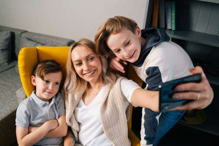 Close-up of happy mother laughing taking selfie with two sons on smartphone sitting in yellow armchair in cozy living room. Smiling bonding family sitting on couch taking selfie using mobile phone.の写真素材