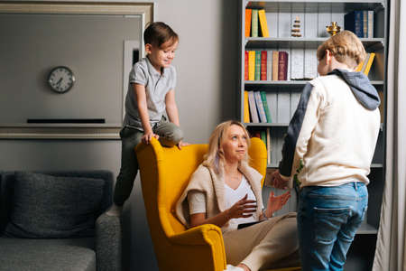 Two loving child son presenting to surprised mom gift box on Mothers Day in cozy living room. Happy cheerful young woman receives gift from loving sons. Concept of family holidays celebration.の写真素材