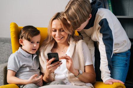 Cheerful young mother with childs having fun using smartphone at home sitting hugs in cozy yellow armchair. Pretty mom with little sons kid in interior of cozy living room looking at screen of phone.の写真素材