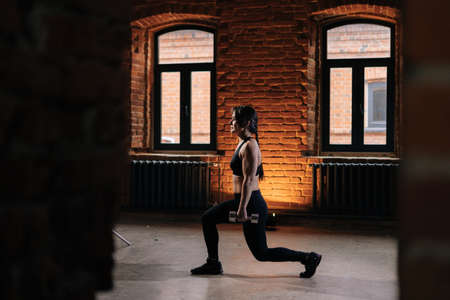 Muscular young athletic woman with beautiful strong body wearing sportswear doing squat and holding dumbbells. Caucasian fitness female workout out exercising in dark gym.の写真素材