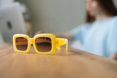 Close-up of stylish yellow sunglasses against background of blurred unrecognizable young woman freelancer using laptop computer sitting at desk. Pretty Caucasian female remote working or studying.の写真素材