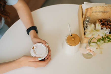 High-angle view of unrecognizable woman holding cup of morning hot coffee sitting at white table in...