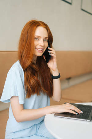 Vertical portrtait of smiling young woman talking on phone and using laptop sitting at table by window in cozy light cafe, looking at camera. Pretty redhead Caucasian lady remote working or studying.の写真素材