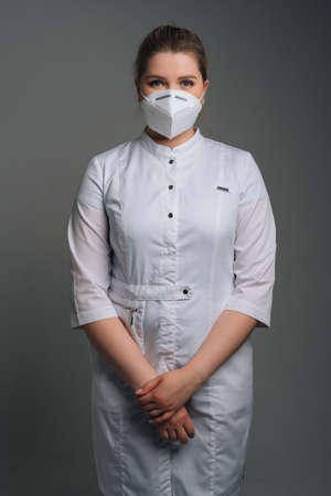 Vertical portrait of young female doctor wearing medical coat and protective face mask standing on dark isolated background. Studio shot of nurse posing looking at camera.の写真素材