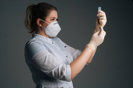 Medium studio shot of nurse in blue gloves and face mask holding and filling up vaccine to syringe on black isolated background. Doctor preparing to give injection of coronavirus vaccine.の写真素材