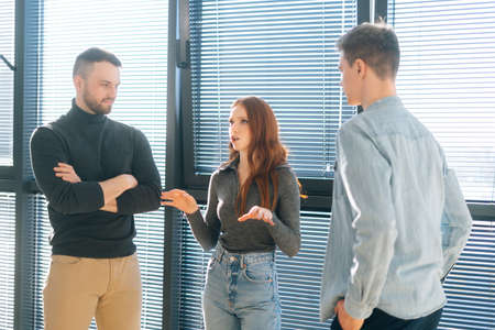 Front view of young redhead businesswoman leader having conversation with male colleagues in modern office room near window. Subordinate office workers listen intently to boss instructions.の写真素材