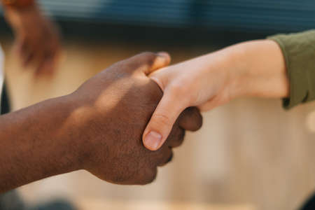 Close-up top view of handshake of Caucasian woman and black male indoors at home office, selective focus. Concept of handshaking black and white hand, interracial friendship and cooperation.の写真素材