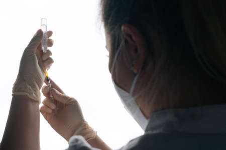 Close-up back view of nurse in white gloves and face mask filling up vaccine to syringe on white isolated background. Studio shot of doctor preparing to give injection of coronavirus vaccine.の写真素材