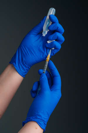 Vertical close-up studio shot of unrecognizable doctor in blue gloves holding and filling up vaccine to syringe on black isolated background. Nurse preparing to give injection of coronavirus vaccine.の写真素材