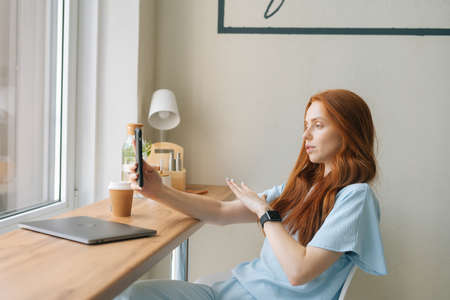 Medium shot of attractive young woman video call on smartphone drinking coffee sitting at table with laptop by window in cafe. Pretty redhead Caucasian lady having leisure activity in coffee shop.の写真素材
