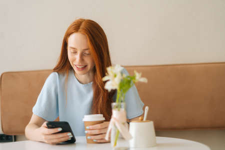 Front view of smiling young woman using smartphone chatting with friends and drinking hot coffee sitting at desk in cozy cafe. Pretty redhead Caucasian lady having leisure activity in coffee shop.の写真素材