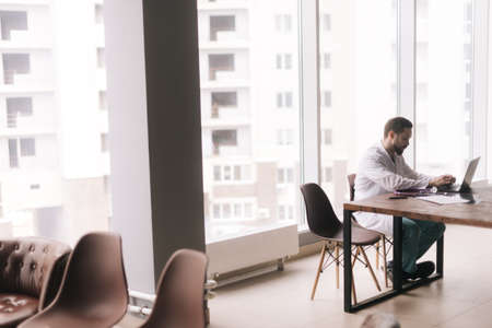 Side view of focused young male doctor wearing white coat working with documents sitting at desk...