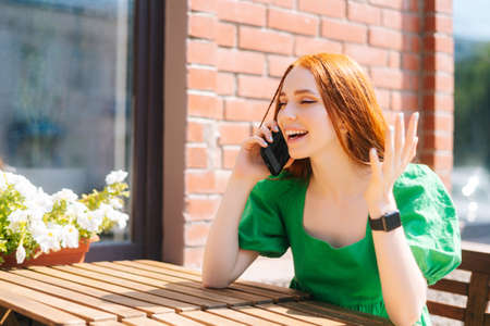 Cheerful young woman having conversation on mobile phone sitting at table in outdoor cafe terrace in sunny summer day. Beautiful redhead lady talking smartphone sitting in coffee shop in European cityの写真素材