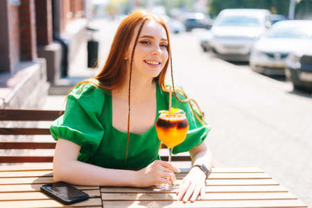 Close-up portrait of happy smiling young woman drinking cocktail through straw sitting at table in outdoor cafe in sunny summer day. Beautiful female student drinking cool lemonade through straw.の写真素材