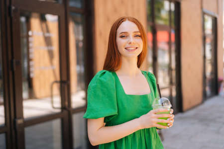 Medium shot portrait of cheerful attractive young woman holding cool cocktail standing at city street in sunny summer day, looking at camera. Pretty lady drinking cold beverage through straw.の写真素材