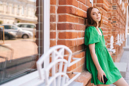 Portrait of sensual young woman holding mobile phone standing leaning on red stone wall of building in town at city street in summer day, looking at camera, blurred background, selective focus.の写真素材