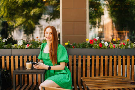 Portrait of smiling redhead young woman holding mobile phone sitting at table with coffee cup in...