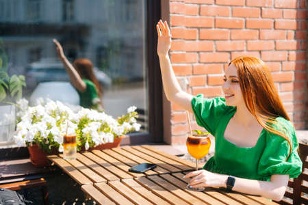 Happy smiling young woman raising hand in sign of salute, greeting someone saying hi with hand gesture sitting at table in outdoor cafe in sunny summer day, drinking cocktail lemonade through straw.の写真素材