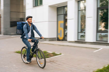 Cheerful handsome young delivery man with thermo backpack riding bicycle in city street on blurred...