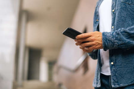 Close-up hands of unrecognizable man texting message on mobile phone outdoors on blurred background, side view, selective focus. Closeup view of male typing sms on modern smartphone outdoors.の写真素材