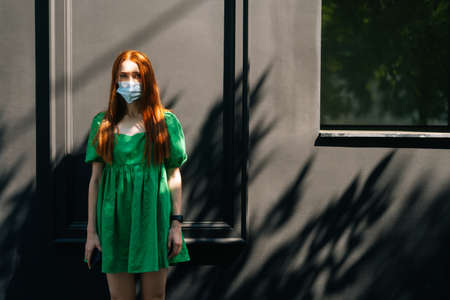 Portrait of confident attractive young woman wearing medical mask standing at city street in sunny summer day, looking at camera, ready for vaccination campaign, dark background.の写真素材