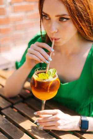 Close-up face of attractive young woman drinking cocktail through straw sitting at table in outdoor cafe in sunny summer day. Beautiful female hipster student drinking cool lemonade through straw.の写真素材