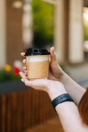 Vertical cropped shot of unrecognizable young woman holding cup with hot coffee sitting at table in outdoor cafe terrace in summer day, blurred background, selective focus, close-up.の写真素材