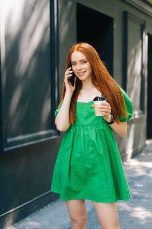 Vertical portrait of smiling young woman in green dress holding takeaway coffee cup, talking on...