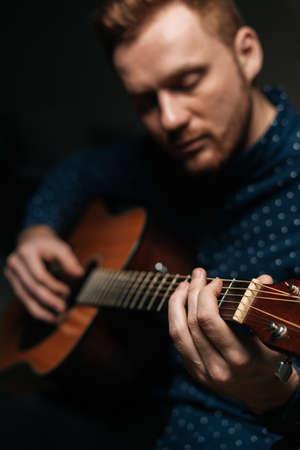 Close-up vertical portrait of guitarist singer male playing acoustic guitar sitting on armchair in dark living room, selective focus. Creative musician enjoying leisure activity in apartment.の写真素材