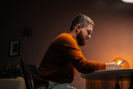 Close-up portrait of handsome young man using typing mobile phone sitting at table in dark living room at home. Closeup of bearded guy using smartphone alone.の写真素材