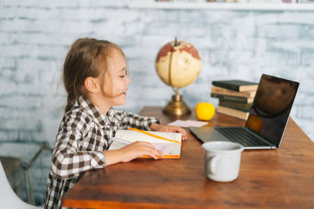 Side view of positive cheerful pupil child school girl doing homework having online lesson and looking at laptop computer screen with charming smile. Concept of online e-learning education.の写真素材