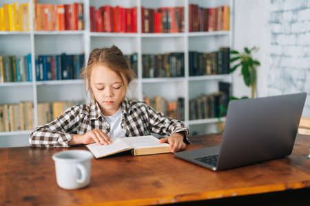 Medium shot portrait of pretty focused primary child school girl reading paper book sitting at table with laptop in cozy children room. Cute primary pupil schoolgirl e-learning online using computer.の写真素材