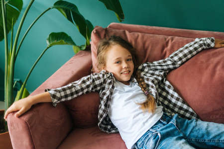 Medium shot of cheerful adorable child kid girl lying on couch with arms outstretched, looking at camera. High-angle view of little cute smiling girl lying on sofa alone in cozy living room.の写真素材