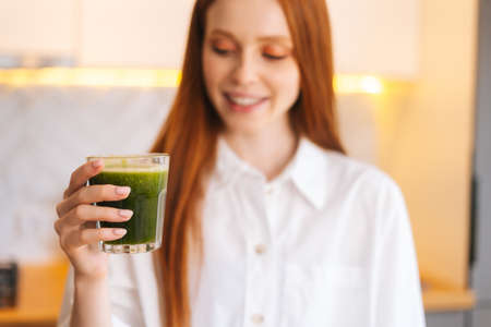 Blurred portrait shot of smiling attractive young woman holding glass with green vegetable detox smoothie cocktail from blender in kitchen. Concept of healthy eating and lifestyle.の写真素材