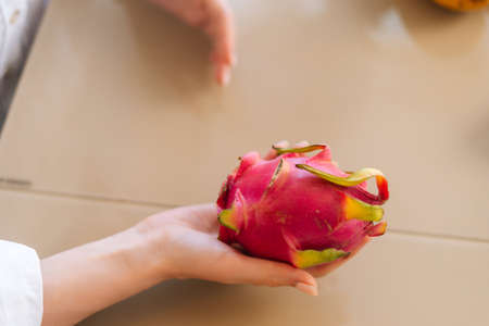 Close-up top view of unrecognizable young woman holding in hands Pitaya dragon fruit sitting at wooden table in kitchen room. Concept of healthy eating and lifestyle.の写真素材