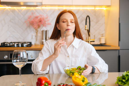 Portrait of attractive happy young redhead woman eating fresh vegetarian salad enjoying fresh tasty vegetables with closed eyes, sitting sitting at table in modern kitchen room, selective focus.の写真素材