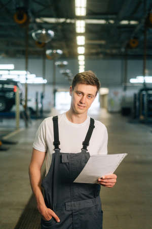Vertical portrait of confident handsome young mechanic male wearing uniform holding clipboard standing in auto repair shop garage, with vehicle background, looking at cameraの写真素材