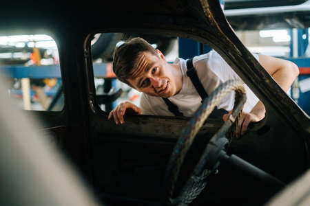 Front view of frustrated young service man in uniform inspecting interior of old car in auto repair shop garage with vehicle background. Concept of car service, repair and maintenance.の写真素材