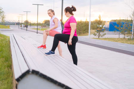 Overweight young woman doing single leg squats using bench with personal trainer outdoor in summer morning. Training from athletic lady for fat female with big abdomen wearing sportswear.の写真素材