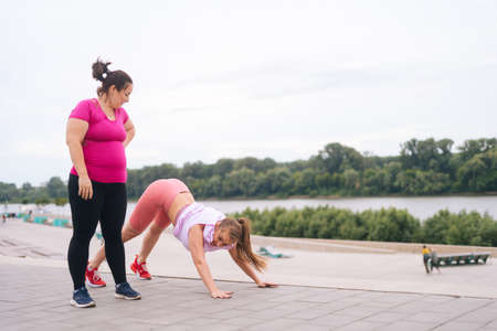 Wide shot of fitness female trainer have personal training with positive overweight young woman outdoor in summer morning. Instructor help fat woman lose weight outside practising yoga asana.の写真素材